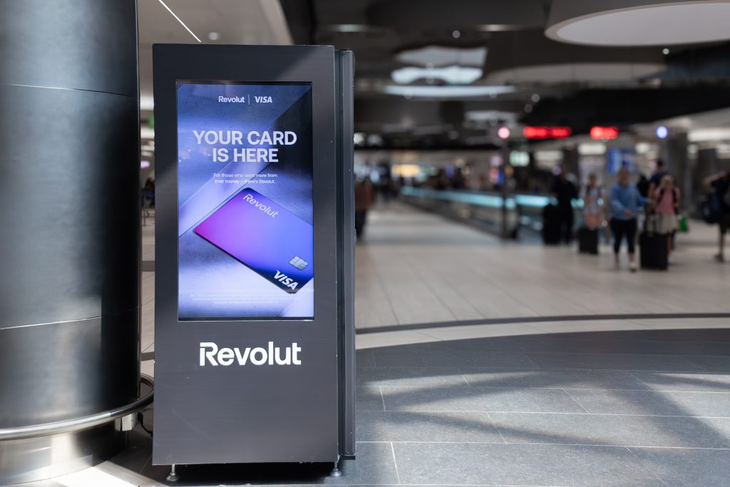 Airport, Fiumicino, Rome,Italy - 30 July 2024: Modern digital signage displaying a vibrant credit card of online bank Revolut advertisement in a bustling airport terminal,